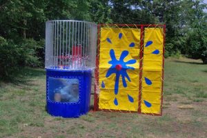 Picture of someone in a dunk tank just as they are falling into the water creating a splash. The Dunk Tank is blue with a window in the base so you can see the person going into the water. The backdrop is yellow and blue with a red target. There are vertical metal bars above the dunk tank to protect the participant while they are sitting on the dunk tank seat from the balls thrown at the target.