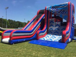 Picture of a ninja warp wall as viewed from the side that has three different wall heights. For those participants who successfully get to the top of their wall their is a slide on the side for them to slide down to the finish area. The inflatable is blue and red with a white slide and a white climbing area.