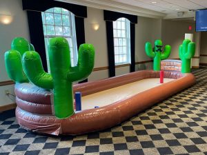 Long, narrow rectangular inflatable arena that has one foot high side walls that are brown. The interior of the arena is not inflated. Their are two green inflatable cacti at either end. Inside the arena are two foam poles to use with oversized foam horseshoes.